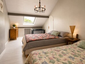 Ein Schlafzimmer mit schrägem Dach, einem Chandelier, einem Oberlicht, einem Bett mit floralem Bettbezug, einem Nachttisch mit Lampe und einem hölzernen Beistelltisch.