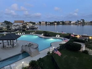 A swimming pool with lounge chairs and American flags, surrounded by a grassy area and a view of a marina and buildings in the background. - Best Views in Orange Beach!! (Orange Beach)