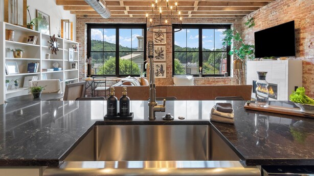 A modern kitchen with a large island, stainless steel sink, and pendant lighting. There is a brick wall and a large window with a view of greenery.