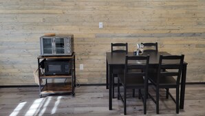 A room with a wooden wall, a white air conditioner, a microwave, a toaster, and a dining table with chairs.