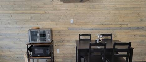 A room with a wooden wall, a table with chairs, a microwave, a toaster, and an air conditioning unit.