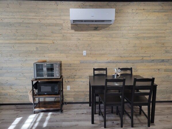 A room with a wooden wall, a table with chairs, a microwave, a toaster, and an air conditioning unit.