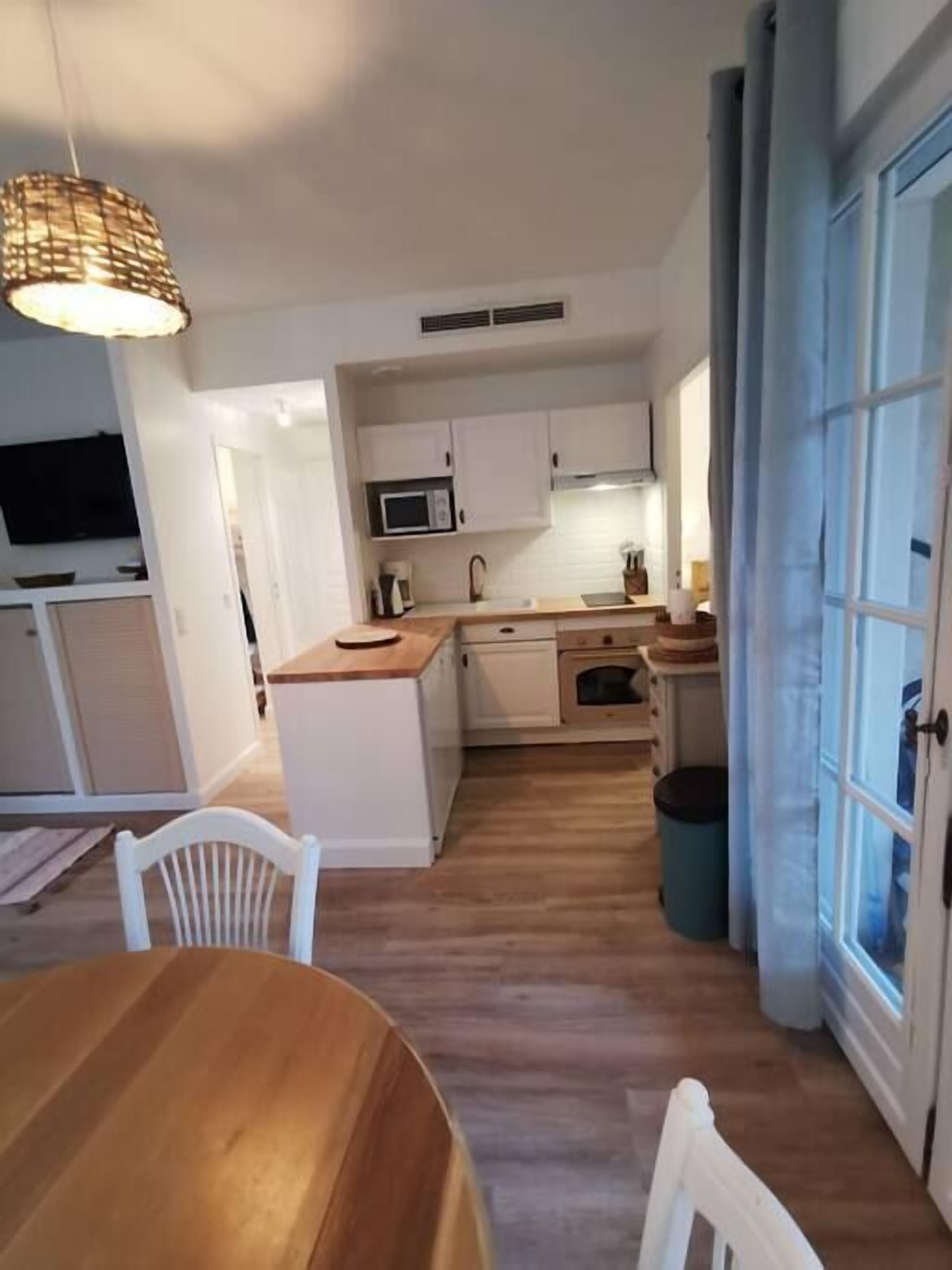 A compact kitchen with white cabinets, a microwave, and a sink. There is a wooden table and chairs in the foreground, and a trash bin is visible.
