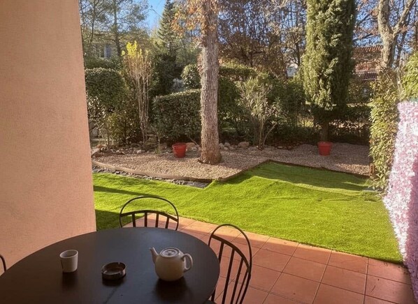 A patio with a table and chairs, a small garden with grass and trees, and a red pot on the ground.