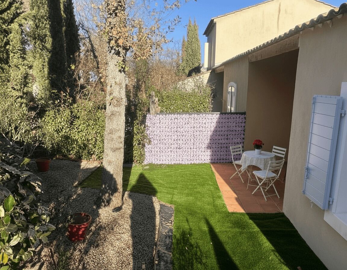 Eine Terrasse mit Tisch und Stühlen, ein kleiner Gartenbereich mit einer Hecke und ein Gebäude mit einem Ziegeldach.