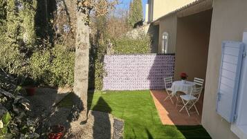 Eine Terrasse mit Tisch und Stühlen, ein kleiner Gartenbereich mit einer Hecke und ein Gebäude mit einem Ziegeldach.