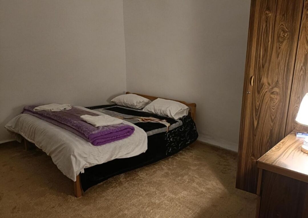 Een kleine kamer met een enkel bed, een houten hoofdeinde, een nachtkastje met een lamp en een vloerbedekking.