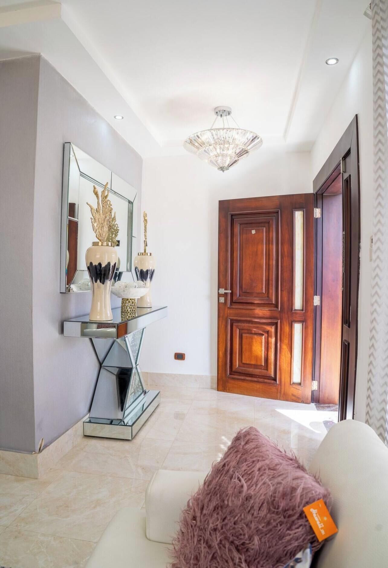 A modern interior with a white sofa, a fluffy pink throw, a mirrored console table with decorative vases, and a wooden door.