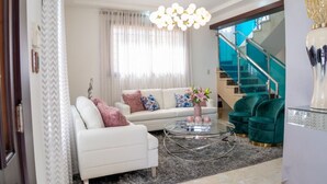 Uma sala de estar moderna com um sofá branco, poltronas verdes, uma mesa de centro de vidro e uma escada com guarda-corpo de vidro.
