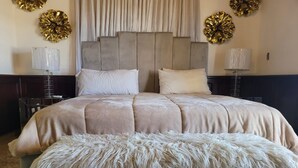 Uma cama grande com cabeceira estofada, duas mesas de cabeceira e um banquinho branco fofo.