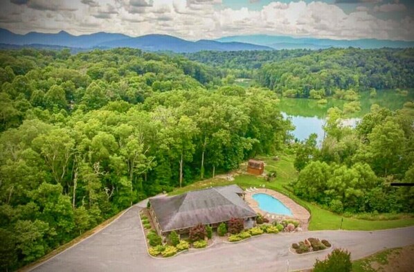 An aerial view of a property with a swimming pool, a large house with a dark roof, and a surrounding forested area.