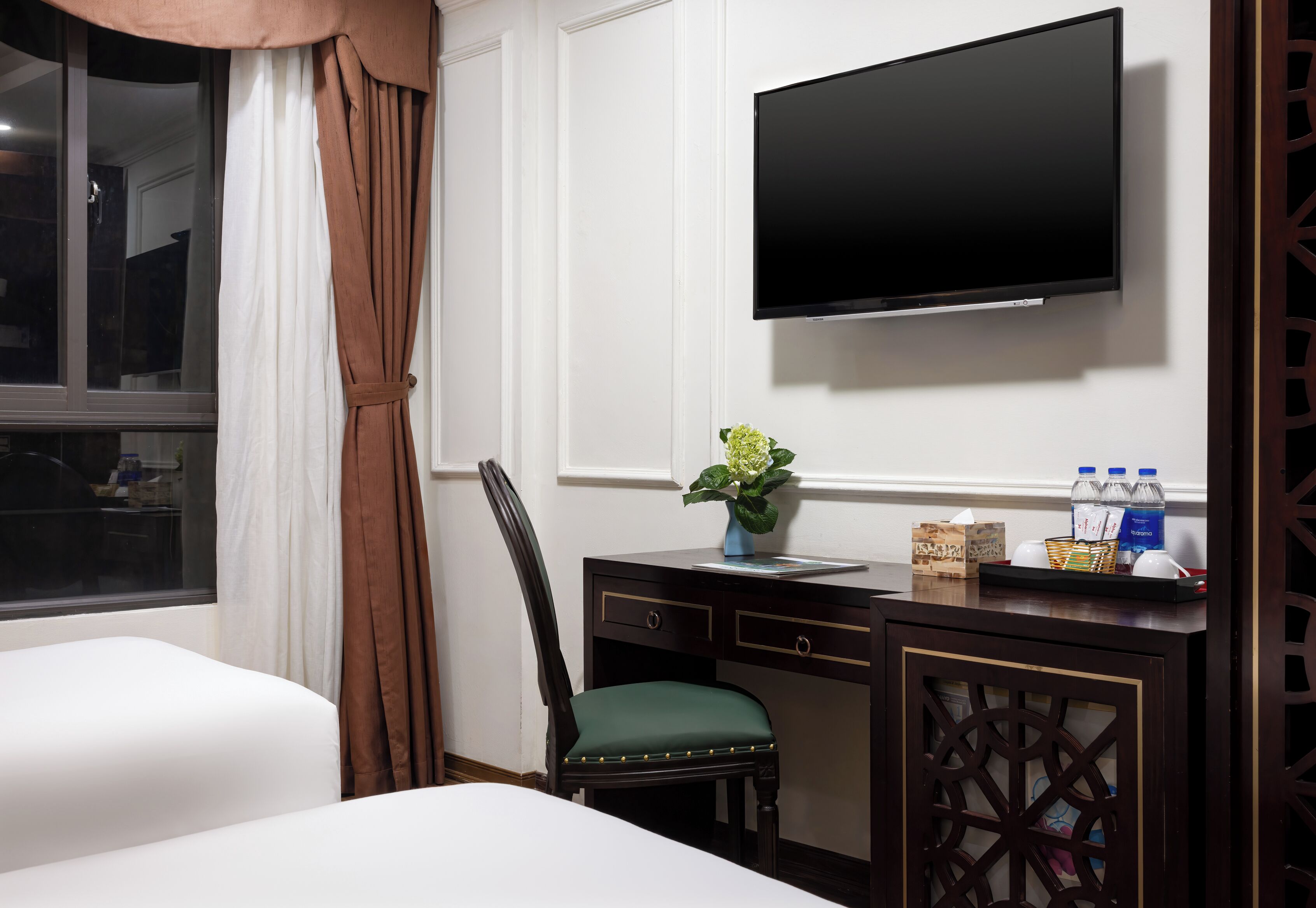 A hotel room with a bed, a desk with a flat-screen TV, a chair, and a window with curtains.