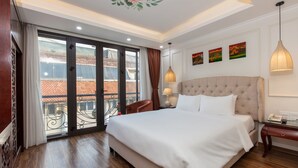 A spacious bedroom with a large bed, a balcony with a view of a red-roofed building, and three paintings on the wall. - Golden Legend Palace Hotel (Hanoi)