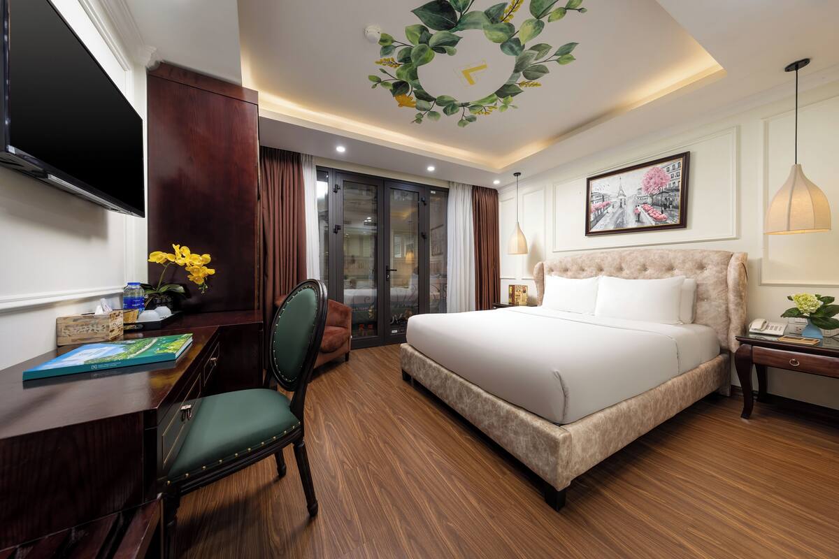 A hotel room with a bed, desk, chair, TV, and artwork on the wall.