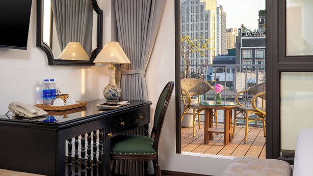 A hotel room with a balcony, a desk with a lamp, a chair, and a view of the cityscape. - Golden Legend Palace Hotel (Hanoi)