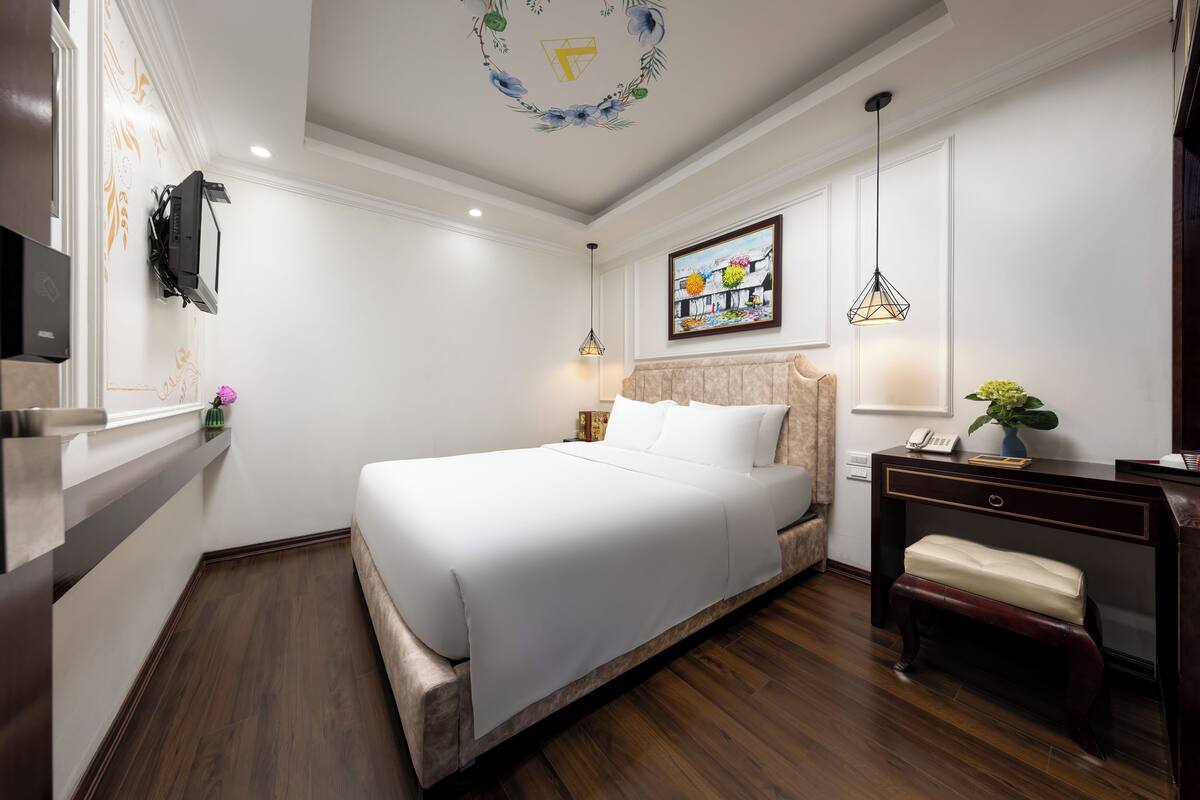 A hotel room with a large bed, a desk with a lamp, a telephone, and a small bench.