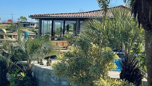 Une maison moderne avec un toit en tuiles, de grandes fenêtres et un patio agrémenté de plantes et de mobilier d’extérieur.