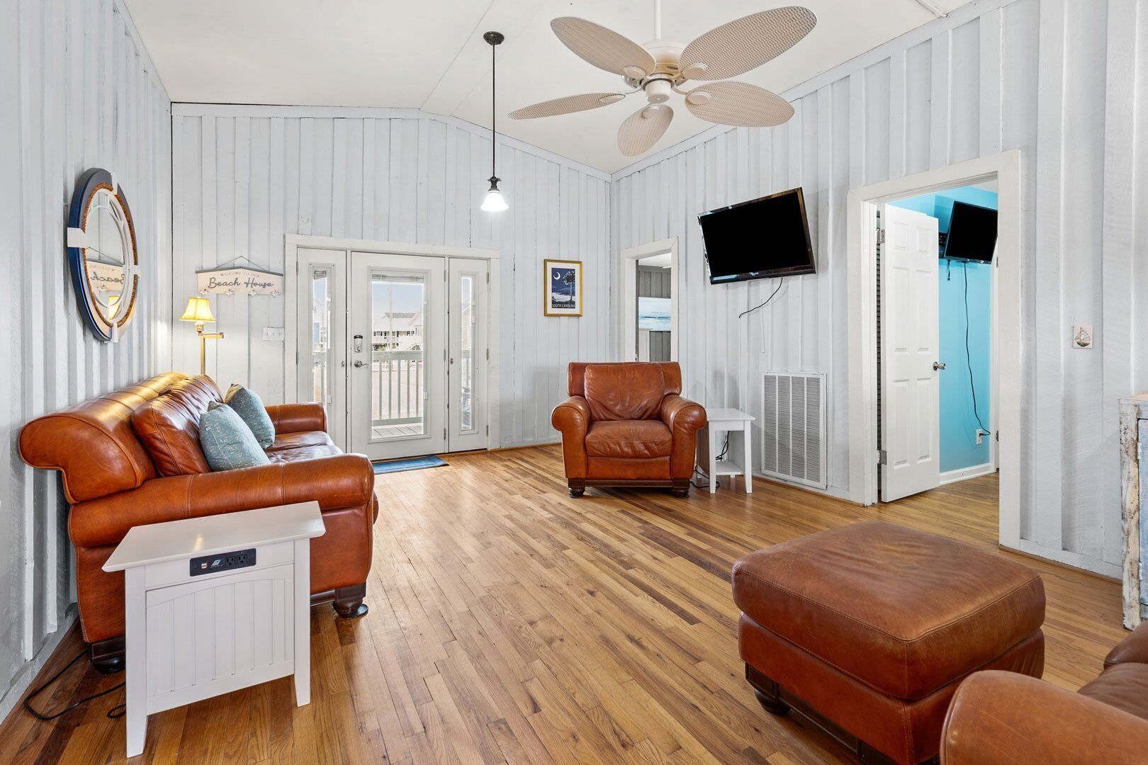 A living room with a brown leather sofa, a brown armchair, and a wooden floor. There is a ceiling fan, a TV mounted on the wall, and a door leading to another room.