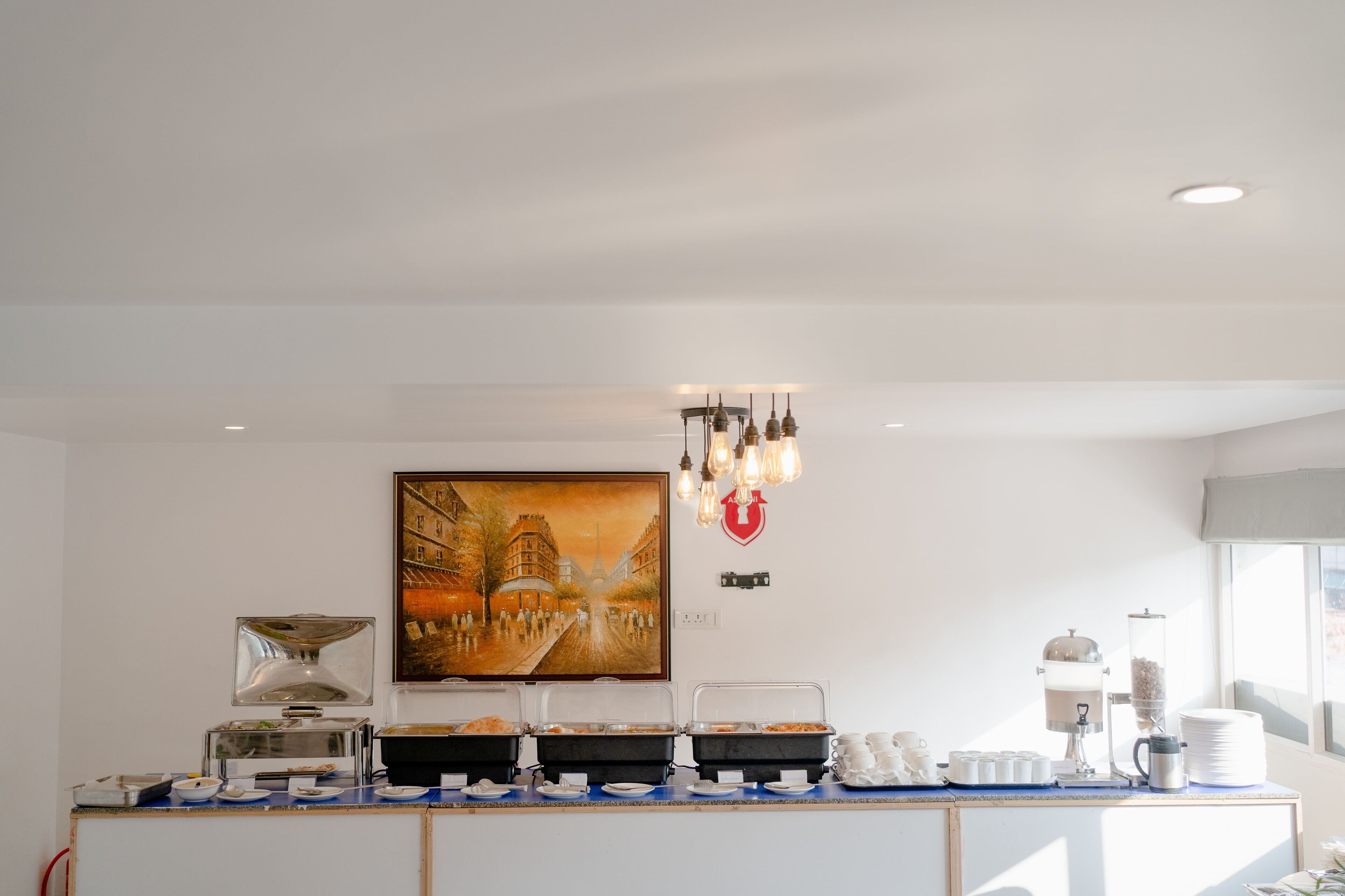 Un buffet pour le petit-déjeuner avec plusieurs points de service, un tableau représentant un paysage urbain et un lustre.