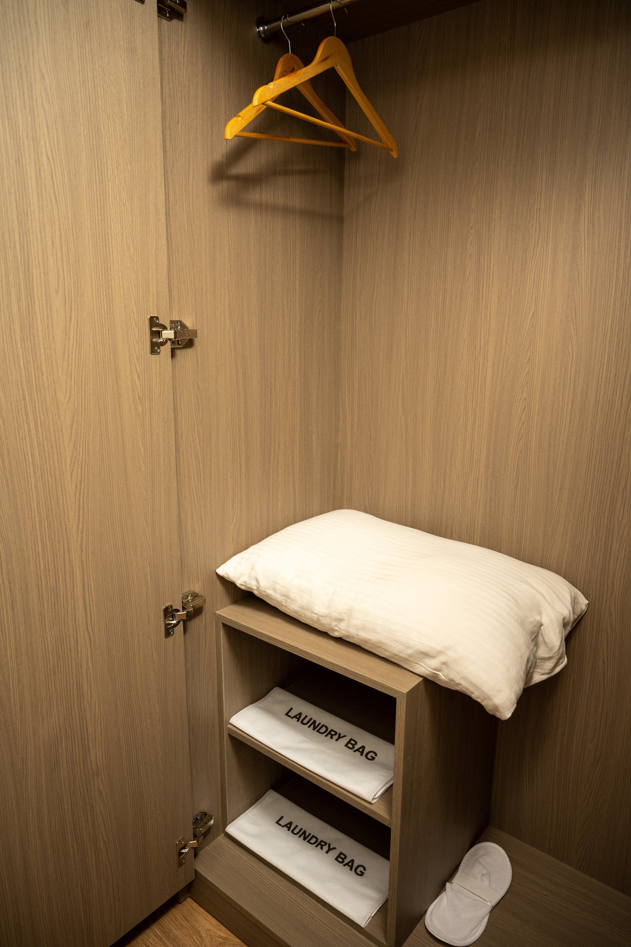 A wooden wardrobe with a hanging rack, two shelves labeled 'Laundry Bag', and a single white pillow on top.