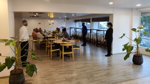 A modern dining area with wooden tables and chairs, potted plants, and a view of the street through large windows. - HOTEL ASRANI INTERNATIONAL (Hyderabad)