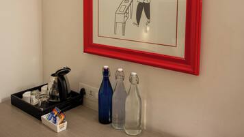 Habitación de hotel con un dibujo enmarcado en la pared, una cafetera, una bandeja con un hervidor, tazas y una botella de agua en la encimera, y un menú en la mesa.