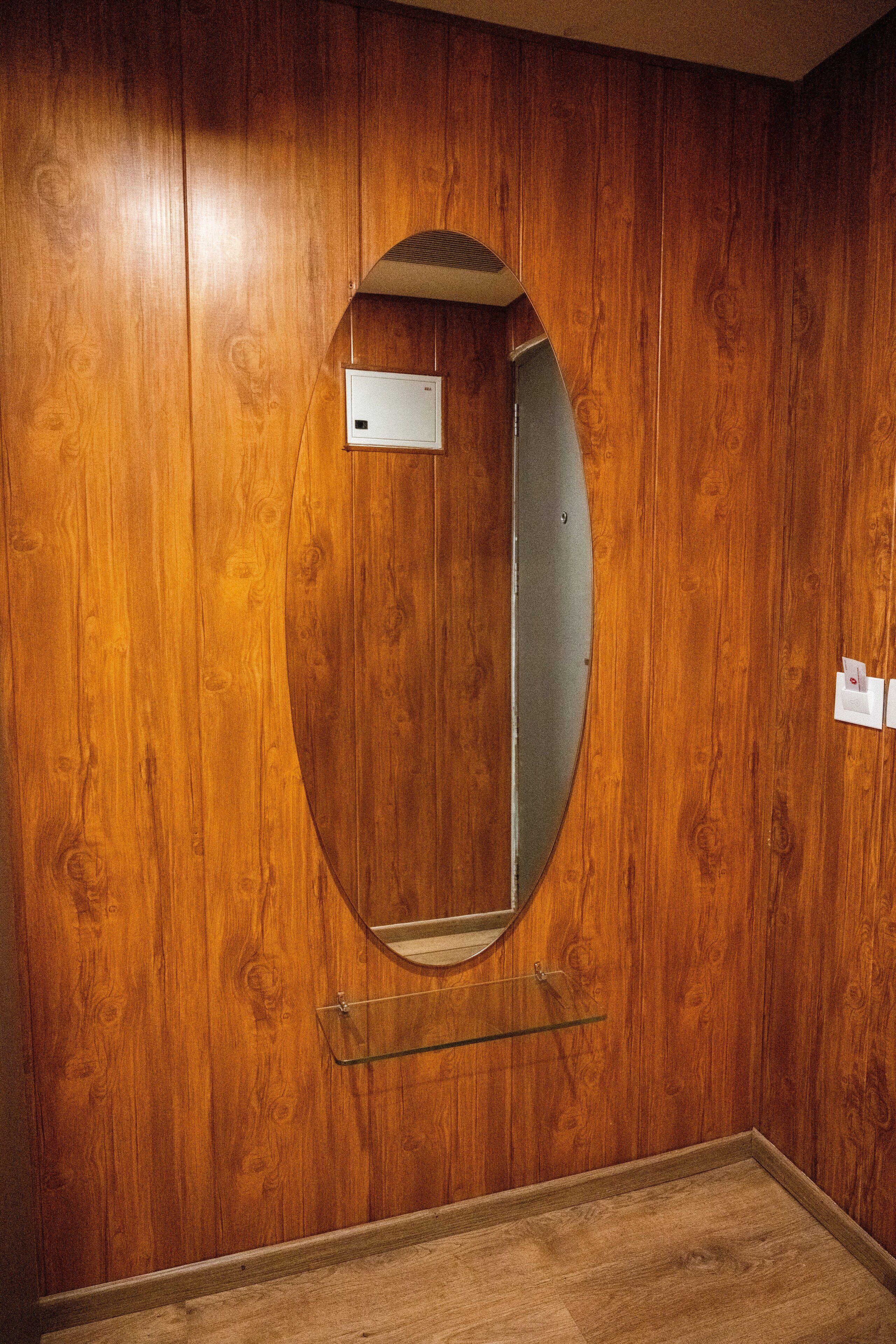 Una habitación con paneles de madera, un espejo circular y una pequeña estantería.