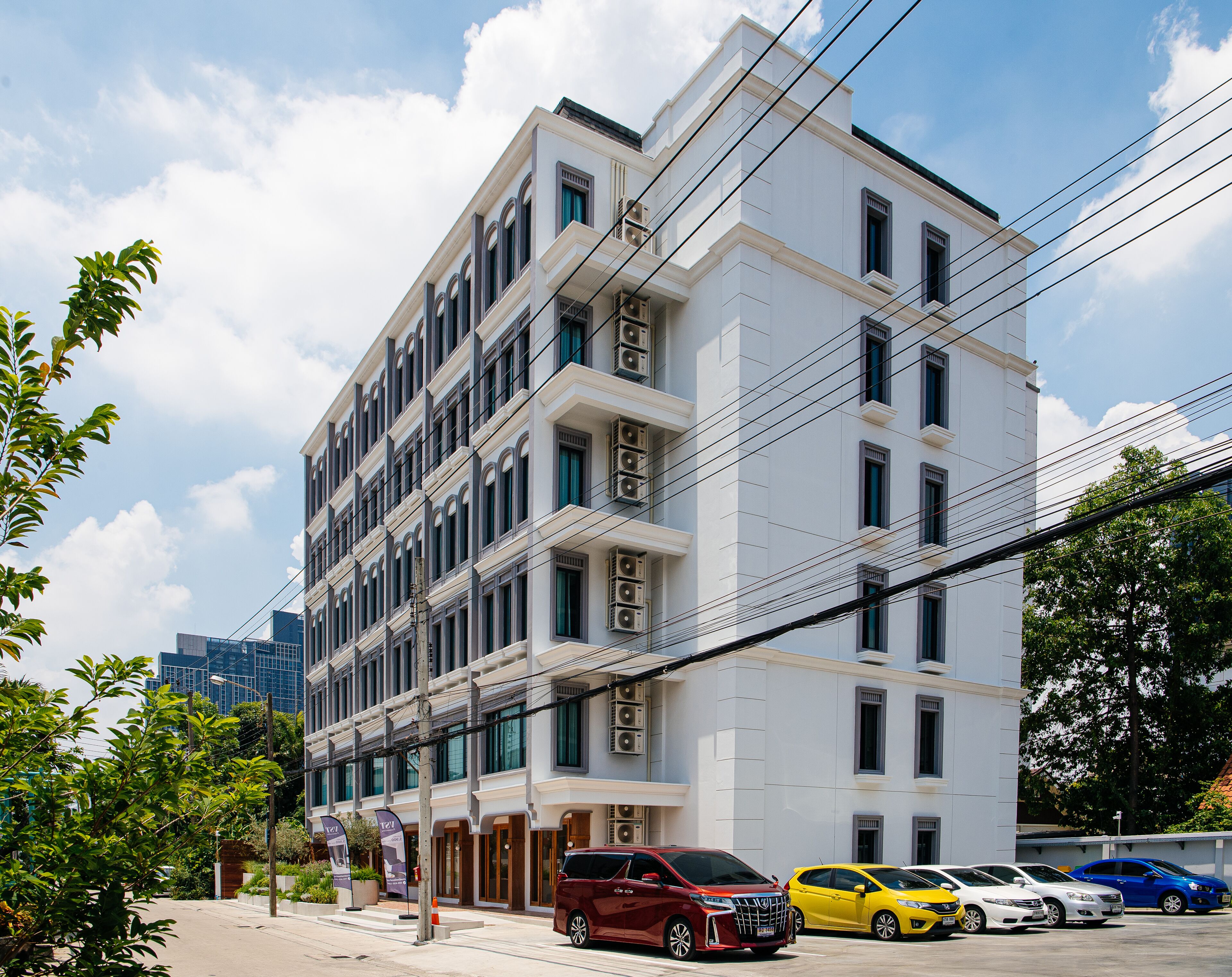 A multi-story building with a white facade, multiple balconies, and a row of parked cars in front.
