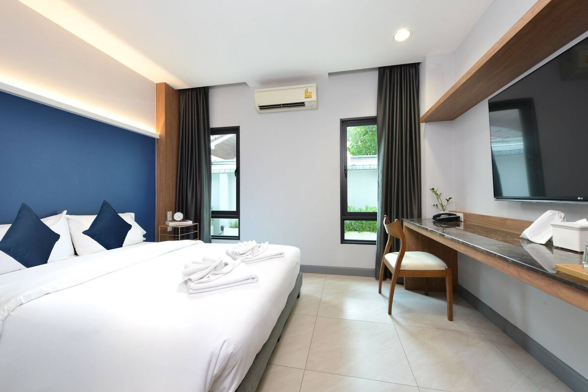 A hotel room with a bed, a desk, a chair, a TV, and an air conditioning unit.