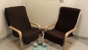 Een kamer met twee bruine fauteuils, een klein tafeltje met twee kopjes en een fles, een kleurrijk vloerkleed, een radiator en een lijst met een afbeelding aan de muur.