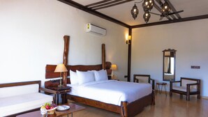 A hotel room with a wooden bed, bedside tables, a small table with fruit, a chair, a mirror, and an air conditioning unit. - Jagira Ananta Elite Gir (Talala)