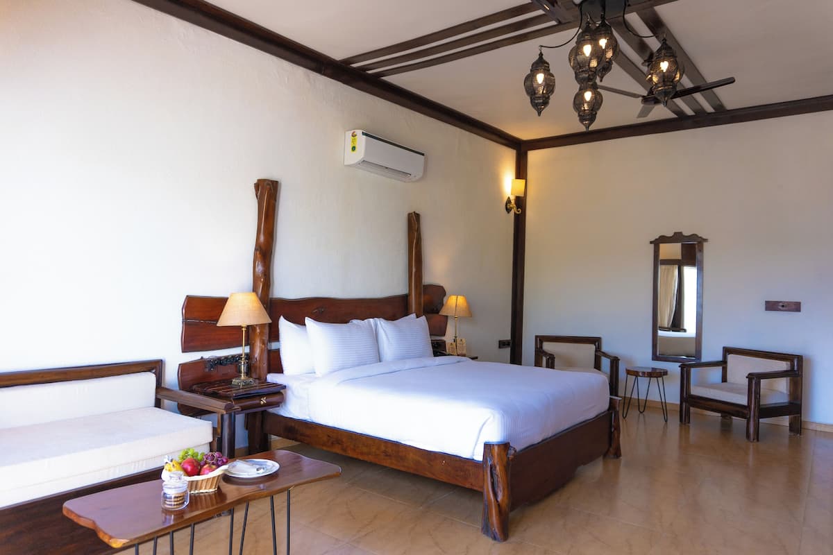 Quarto de hotel com cama de madeira, mesas de cabeceira, uma pequena mesa com frutas, uma cadeira, espelho e unidade de ar condicionado.