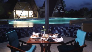 An outdoor dining area with a pool view, a table set with plates, glasses, and a vase with a flower. - Jagira Ananta Elite Gir (Talala)