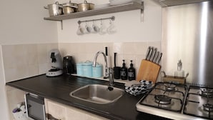 Et køkken med en stålvask, kaffemaskine, elkedel og komfur. Der er skabe under bordpladen og en hylde med gryder og pander.