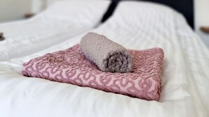 En pænt redt seng med hvide og lyserøde sengetæger, et sammenrullet håndklæde og et maleri af en strand.