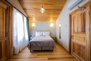 A bedroom with a wooden floor, a bed with white bedding, a nightstand, a wall-mounted lamp, and a large wooden door.