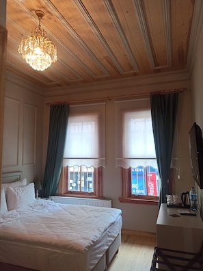 Une chambre d’hôtel avec un plafond en bois, un lit avec du linge de maison blanc, deux fenêtres avec des rideaux, un radiateur et un bureau avec un miroir et une bouteille d’eau.