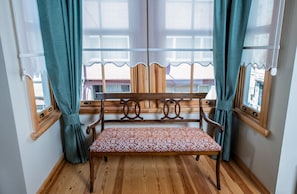 Un banco de madera con un cojín estampado en una habitación con suelo de madera, una ventana con cortinas y vistas al exterior.