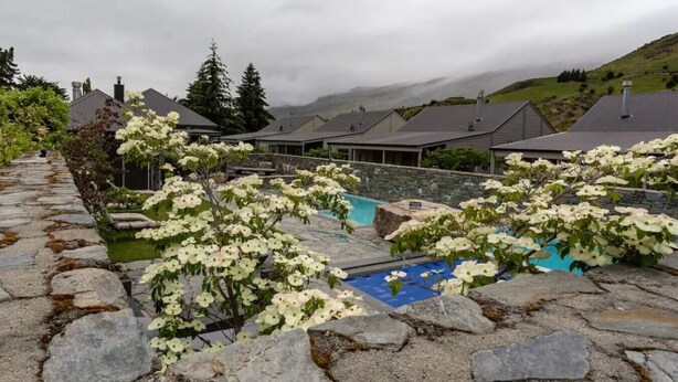 A stone-paved area with a swimming pool, surrounded by white flowering shrubs and a stone wall. - Studio 26 - One Bedroom Place with Pool (Cardrona)