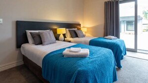 A hotel room with two beds, each with a blue blanket and pillows, a dark headboard, bedside lamps, and a view of the outdoors through a large window. - Studio 26 - One Bedroom Place with Pool (Cardrona)