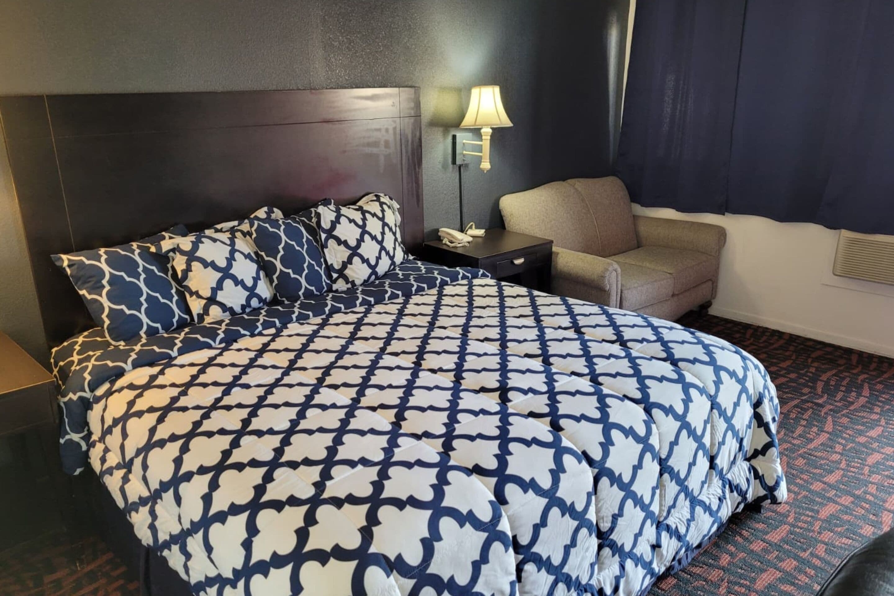 Una habitación de hotel con una cama grande, una mesita de noche con lámpara, una silla y una ventana con cortinas.