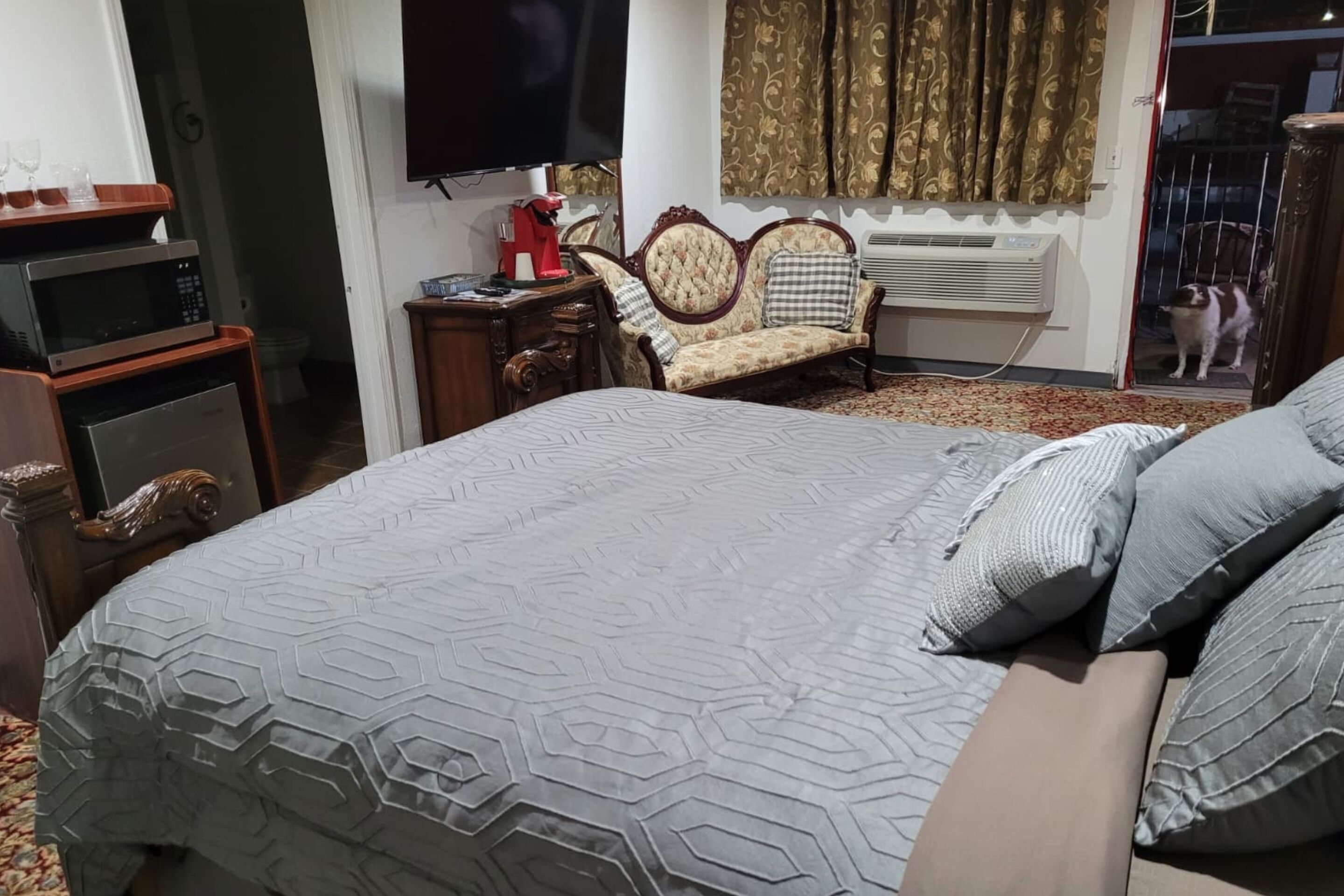 A bedroom with a large bed, a television, a sofa, and a cat in the doorway.
