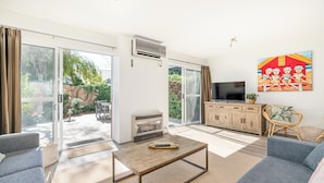A living room with a sofa, a coffee table, a TV, and a sliding glass door leading to a patio. - Broadwater Beach Villa | Pool, Spa & Tennis (Broadwater)