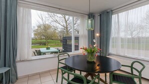 A conservatory with a table and chairs, a vase with flowers, and a view of a park. - 4-Person Stay at Rustenpolder (Vrouwenpolder)