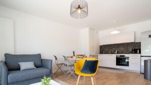 A modern living room with a grey sofa, a white coffee table, and a yellow chair. A kitchen with white cabinets and a black backsplash is visible in the background. - Hemisphère by Interhome (Carnac)