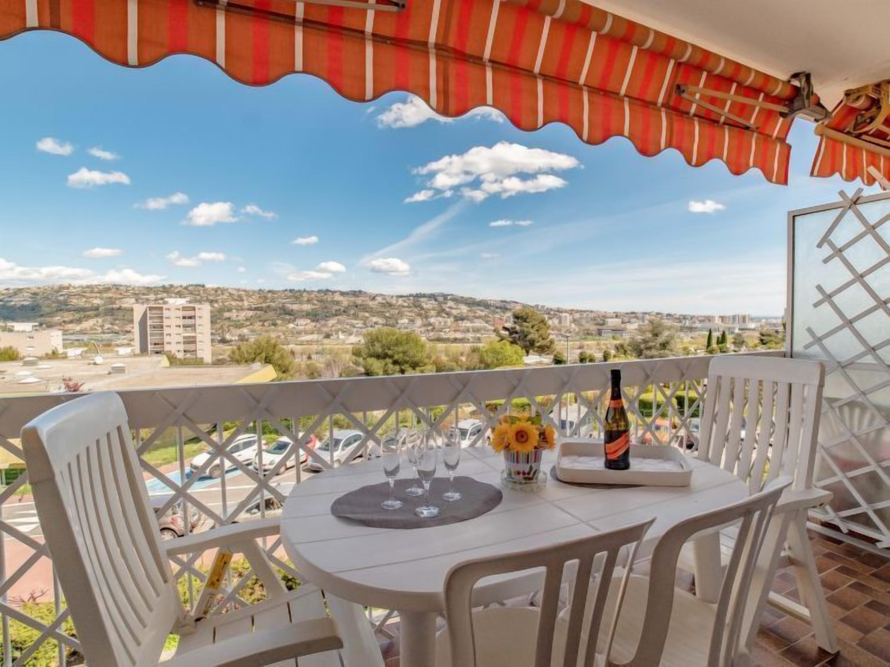 A balcony with a table set for two, a bottle, and a vase with flowers.