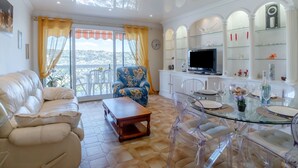 A living room with a sofa, a coffee table, and a dining area with a glass table and chairs. There is a TV on a cabinet, and a view of the city through large windows. - Le Giono by Interhome (Saint Laurent du Var)