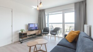 Una sala de estar moderna con un sofá gris, dos mesas auxiliares de madera, un televisor sobre un mueble de madera y un balcón con sillas.
