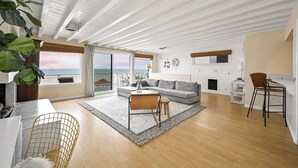 Een ruime woonkamer met een grote bank, een salontafel en een eethoek met een stoel. Door grote ramen is er uitzicht op de oceaan.