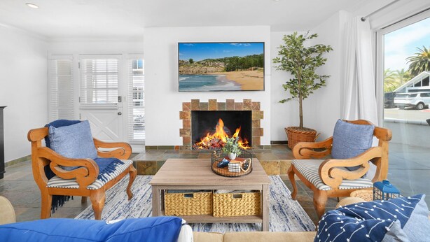 A living room with a fireplace, two wooden chairs, a coffee table, and a large window with a beach view. - Spacious 3 Bedroom Home w/ Fire Pit at the Beach! (Newport Beach)